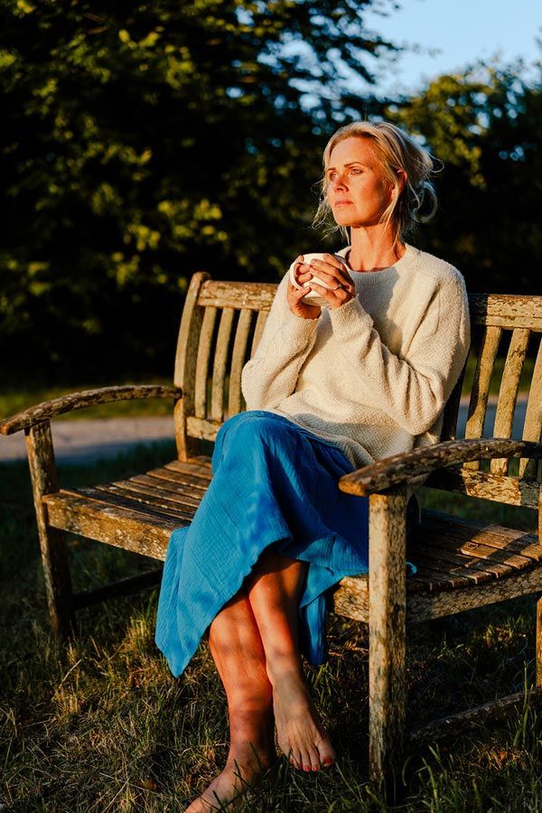 Woman sitting on wooden bench, holding a cup, eyes closed in the sunlight. Blue skirt, cream sweater. Outdoor setting.