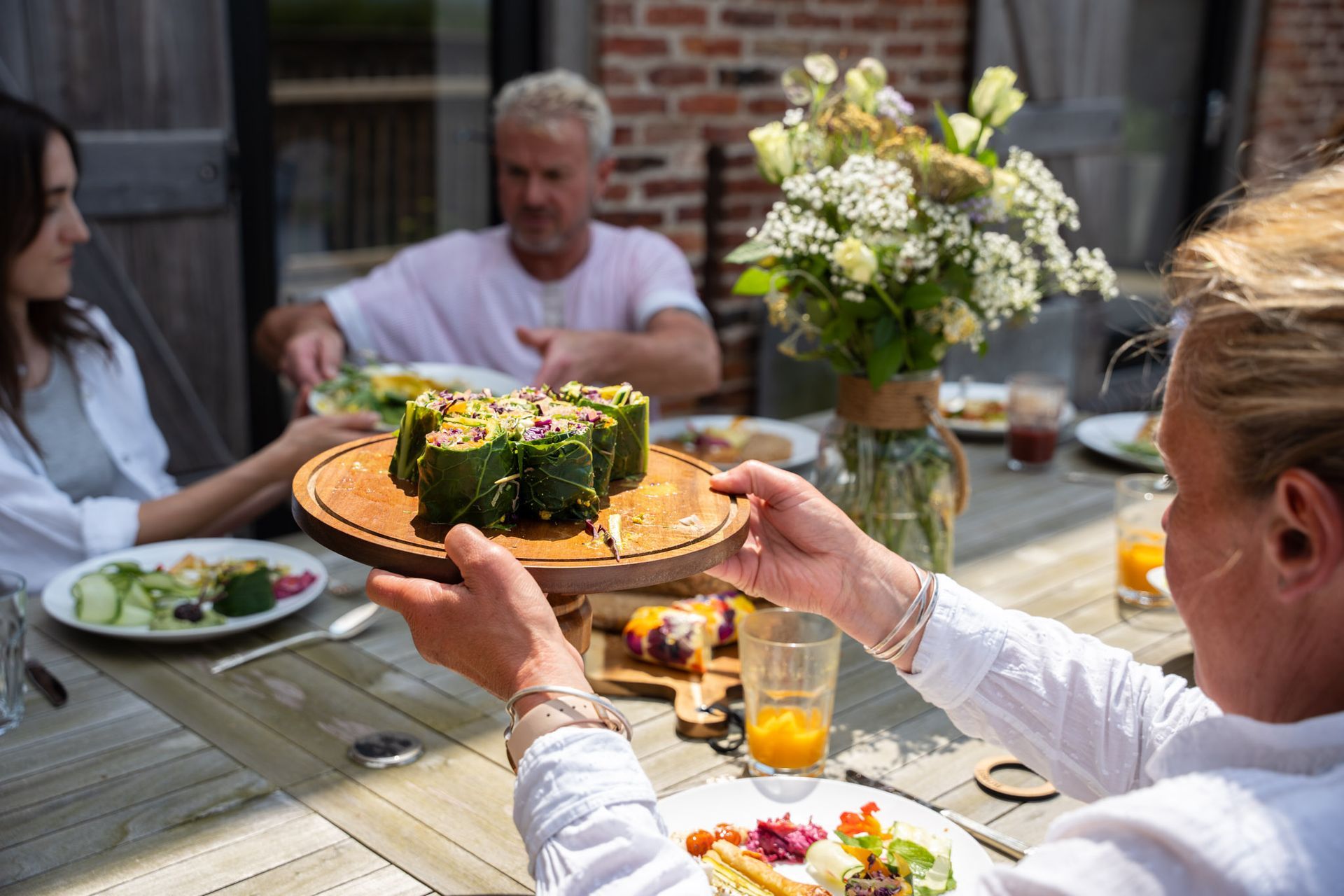 Woman serving stuffed zucchini rolls at outdoor dining table. Two people in background.