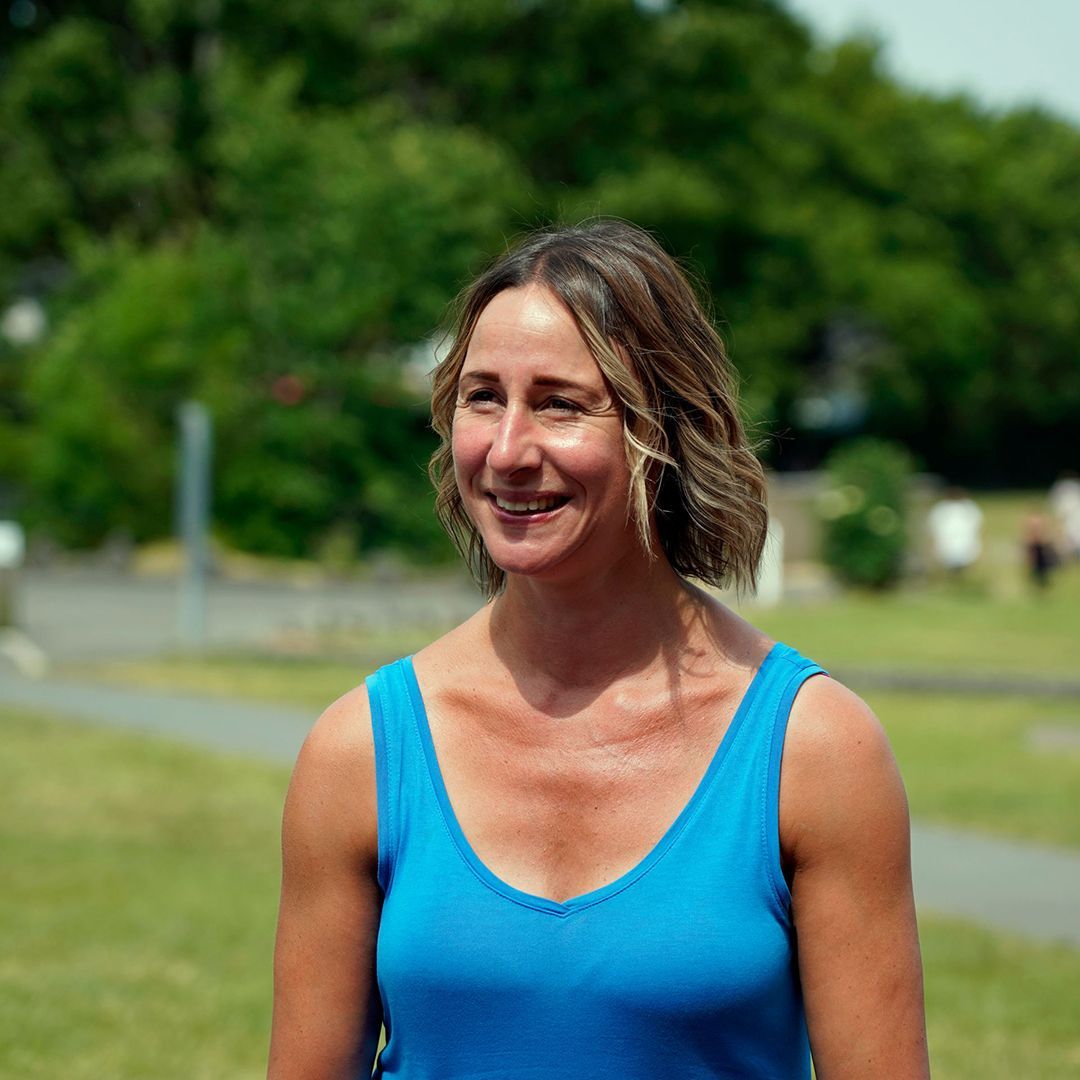 Woman in blue tank top smiling outdoors, with green trees and a park in the background.
