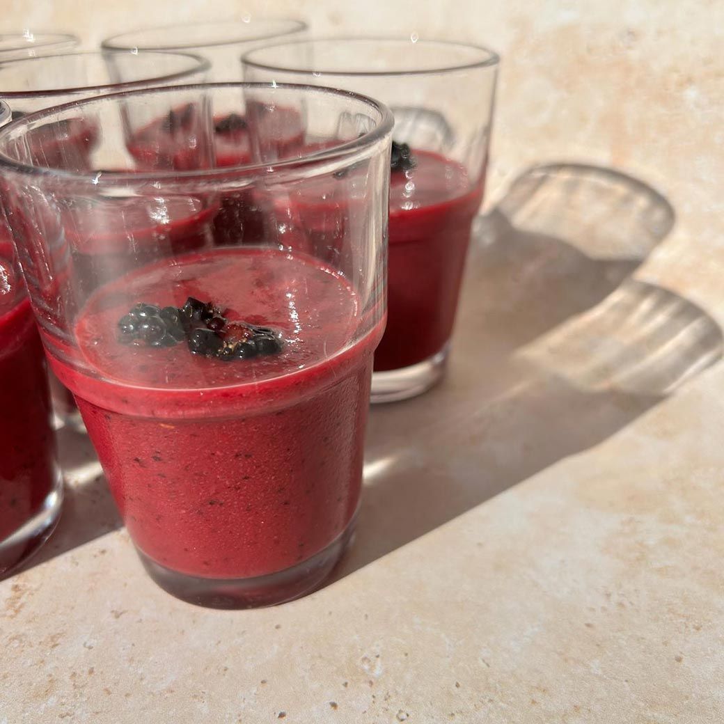 Glasses of red smoothie topped with dark berries, on a light-colored surface with shadows.