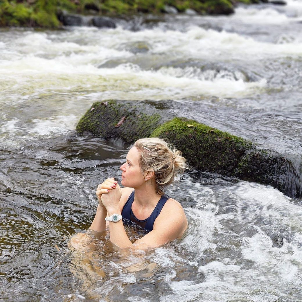 Woman sits in a rushing river, arms crossed, wearing a swimsuit and watch. Water surrounds her in a rocky stream.
