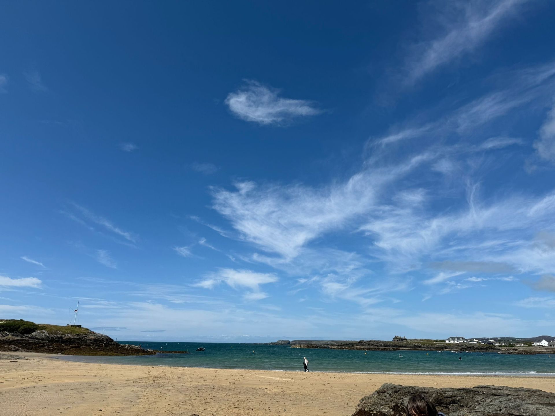A sandy beach meets turquoise water under a vibrant blue sky with wispy clouds, with a lone person walking near the surf.