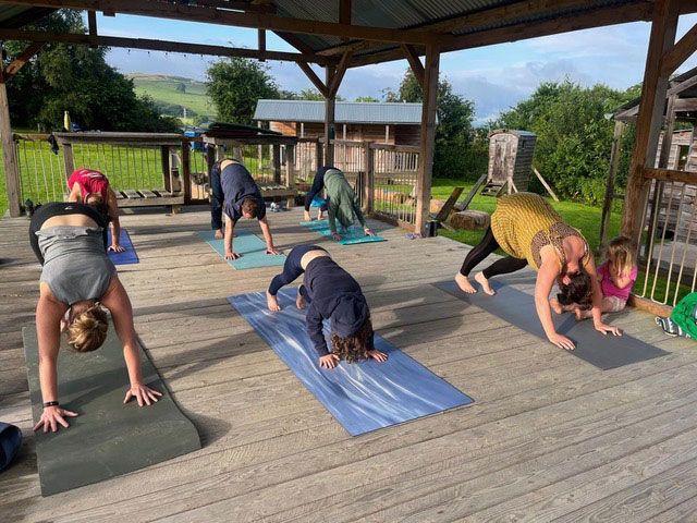 People doing yoga outdoors on a wooden deck under a shelter, overlooking a rural landscape.