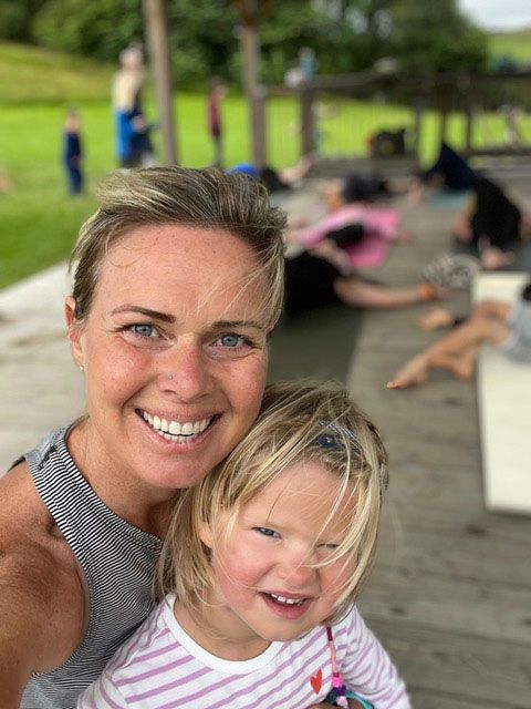 Woman and young child smiling, posing for a selfie. Background: people practicing yoga outdoors.