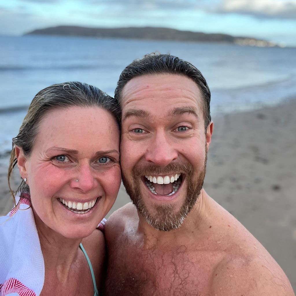 Smiling couple on a beach, woman with towel, man shirtless, ocean in background.