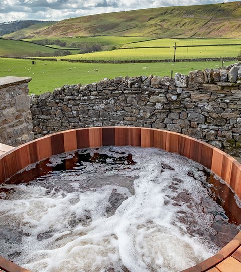 Wooden hot tub with bubbling water, set against a green field and stone wall, rolling hills in the background.