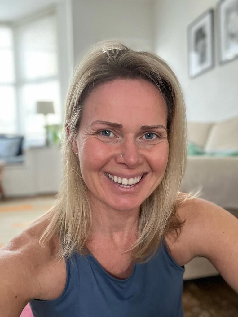 Woman smiling, wearing a blue tank top, indoors with window and sofa in the background.