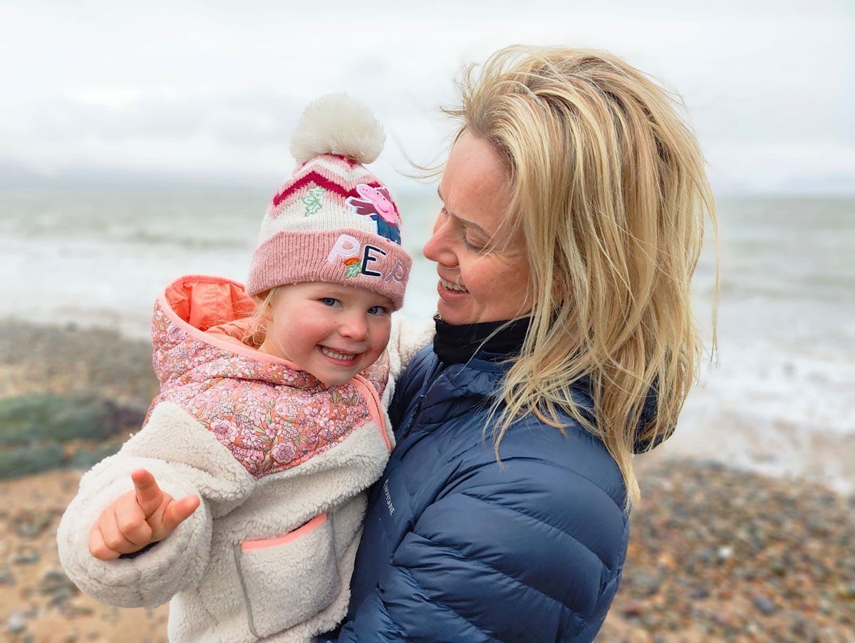 Woman holding a smiling child, both bundled in winter clothing near a beach.