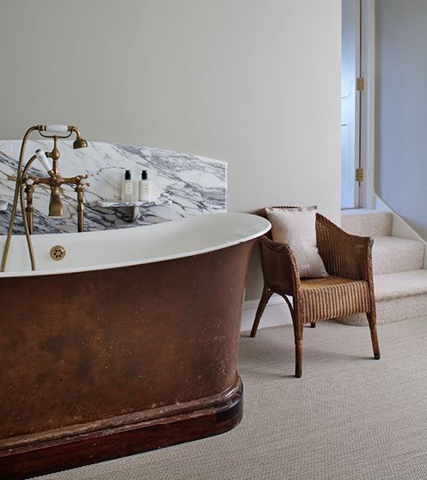 A copper bathtub with a marble backsplash and gold fixtures beside a wicker chair on a carpeted floor.