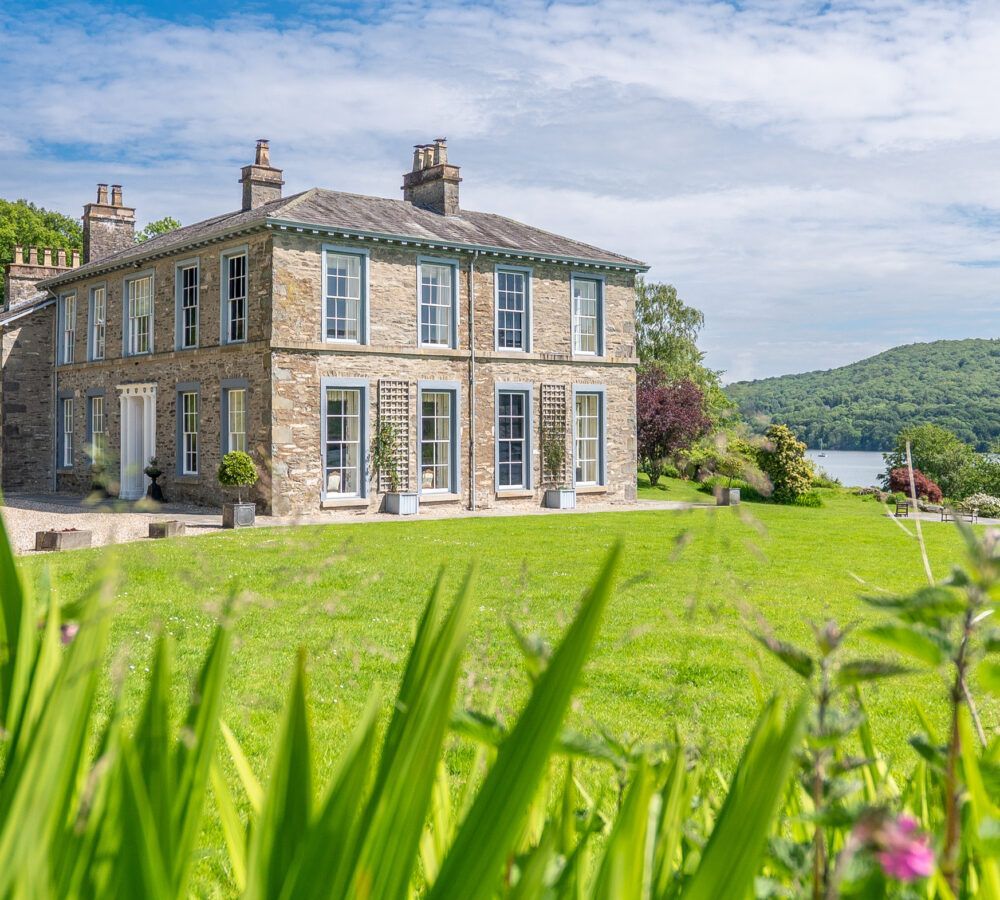 Stone house with green lawn overlooking a lake under a bright blue sky.