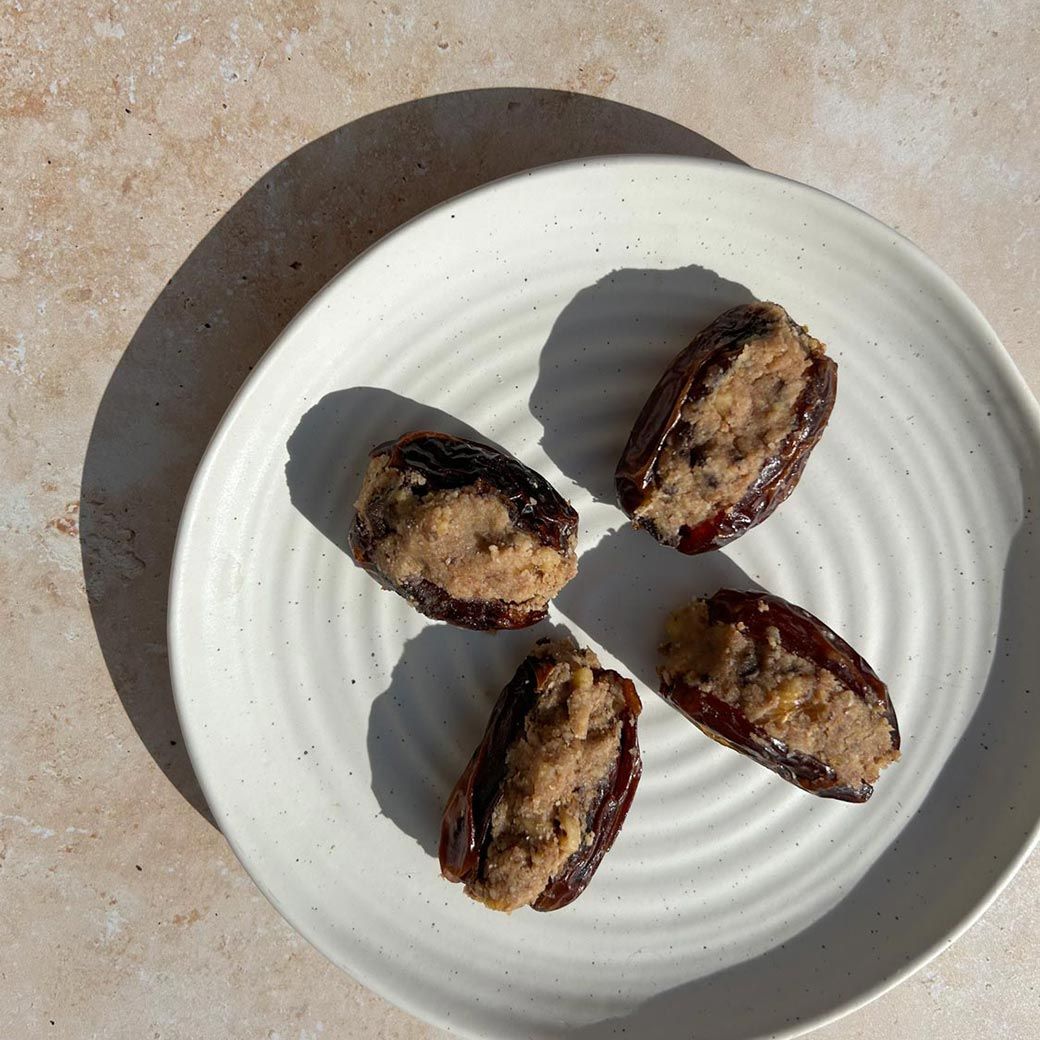 Four dates stuffed with a light-colored filling on a white plate, set on a beige surface with shadows.