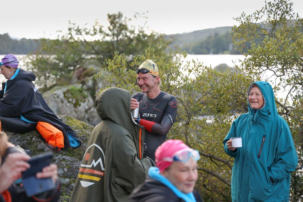 People in swim gear and robes by a lake, some smiling and holding mugs.