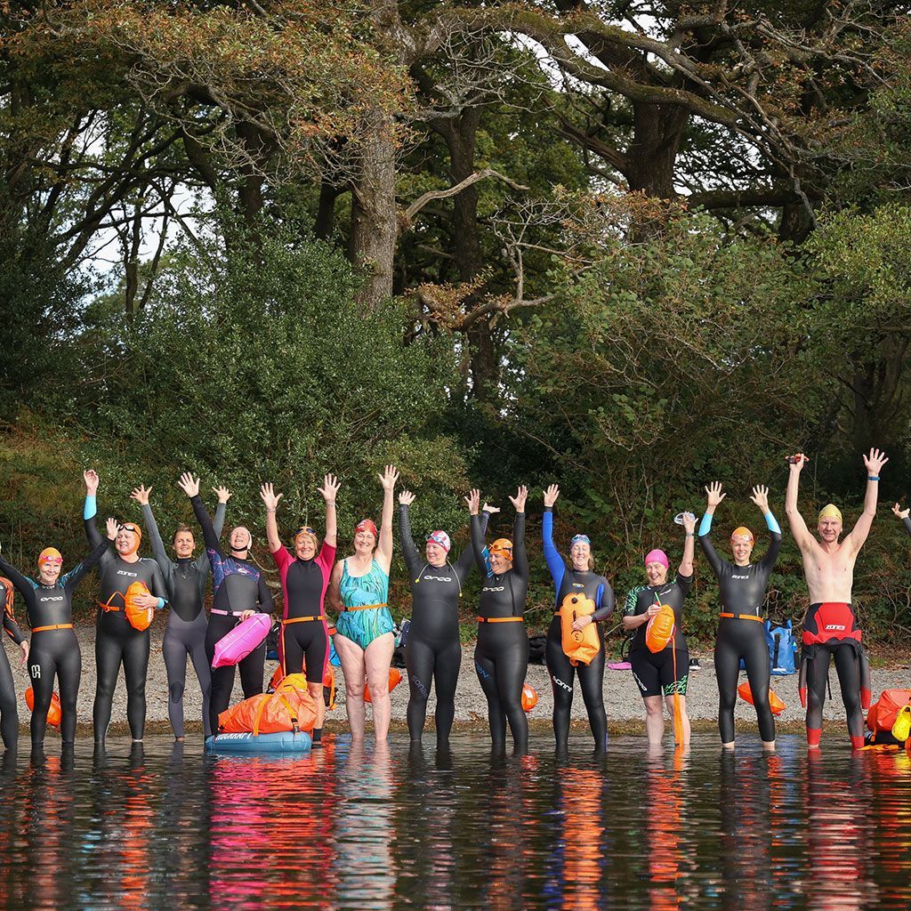 Group of people in wetsuits raise arms on a lake shore, trees in the background.