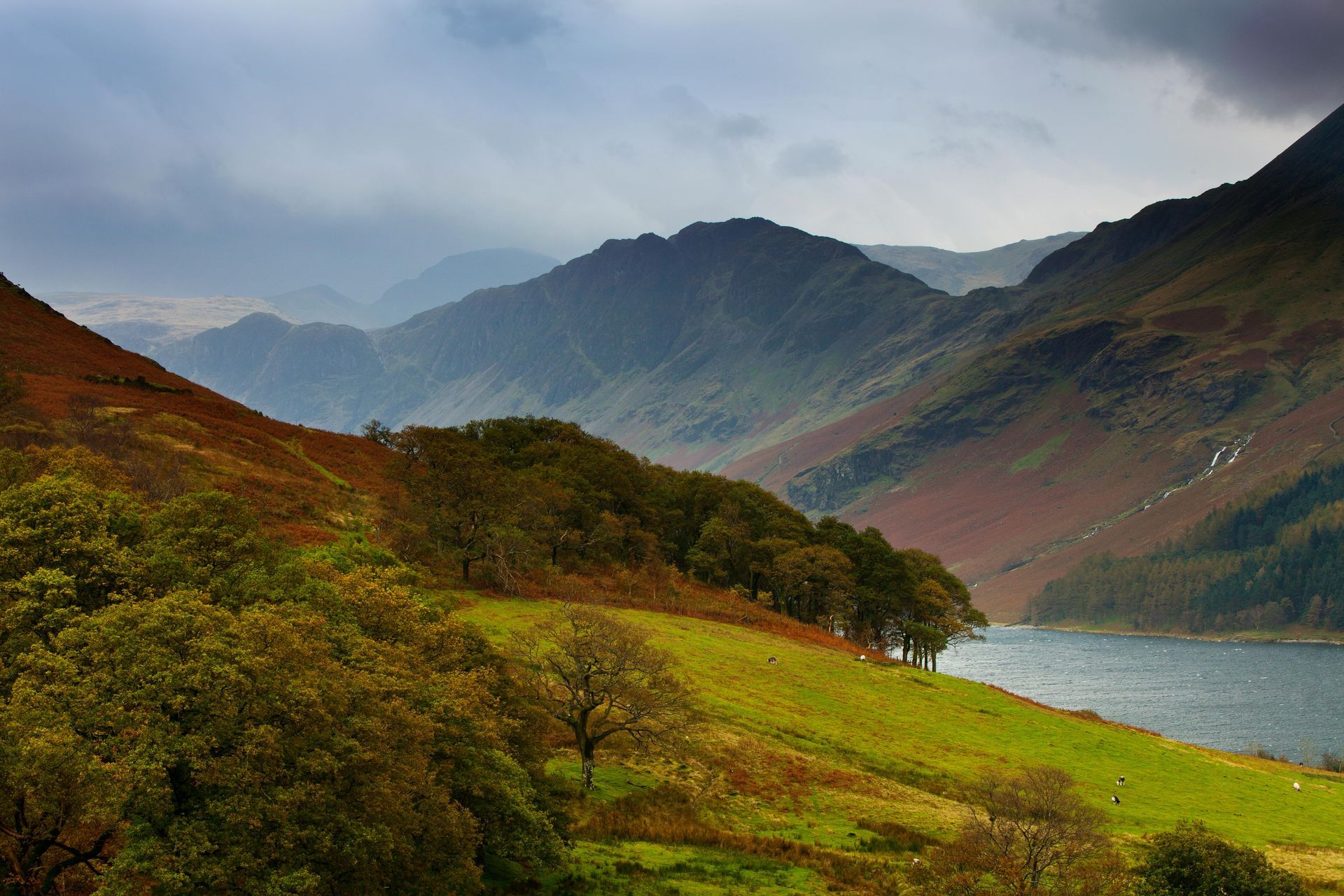 Green hillside descends to a lake. Mountains in background under cloudy sky.