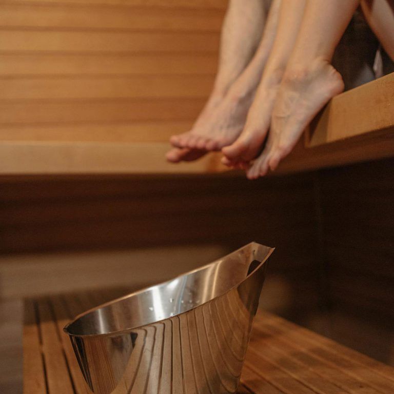 Feet resting on a wooden sauna bench; stainless steel bucket in the foreground.