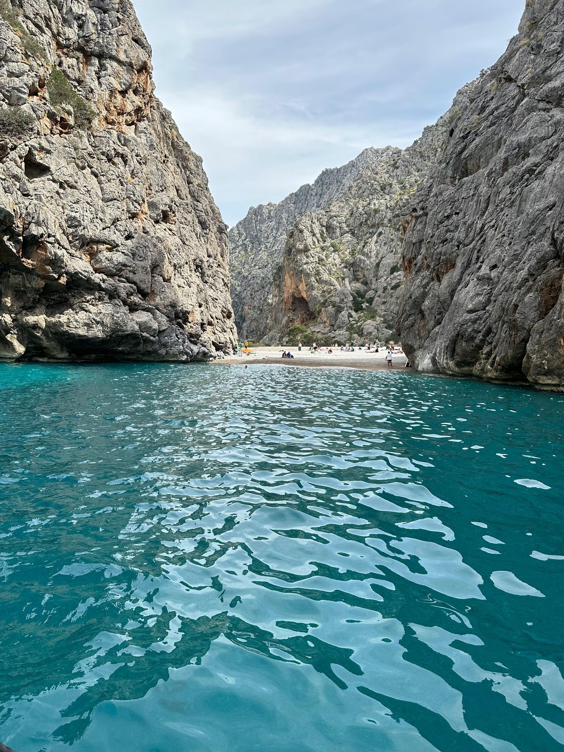 Azure water between towering rock cliffs leads to a small beach under a cloudy sky.