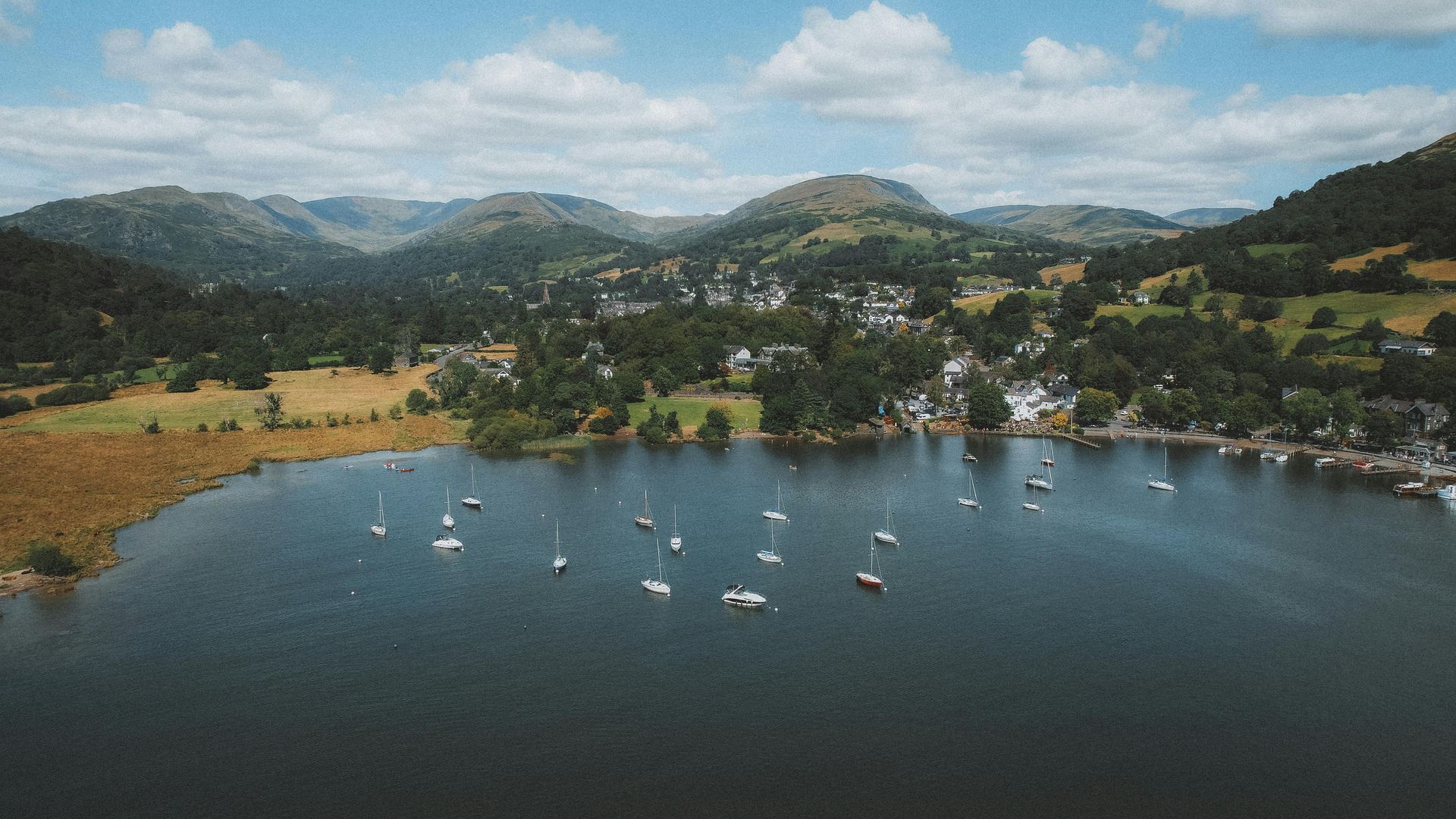 Aerial view of a lake with sailboats, a town, and green hills under a partly cloudy sky.
