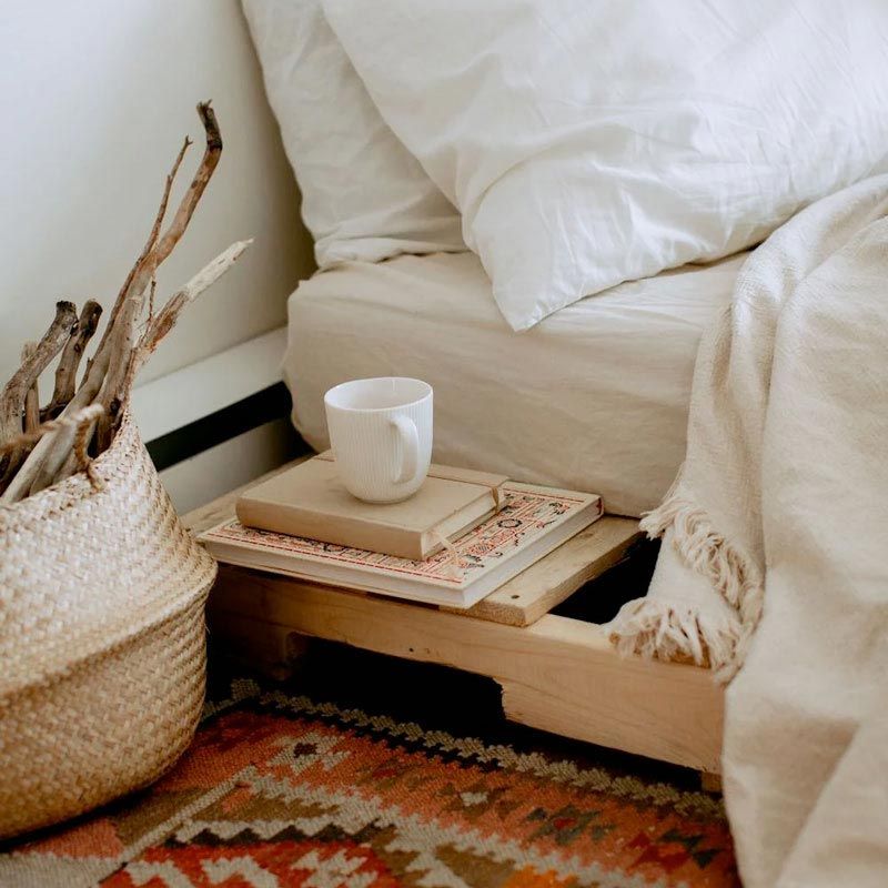 Bedside table with coffee cup, books, blanket, and woven basket of sticks.