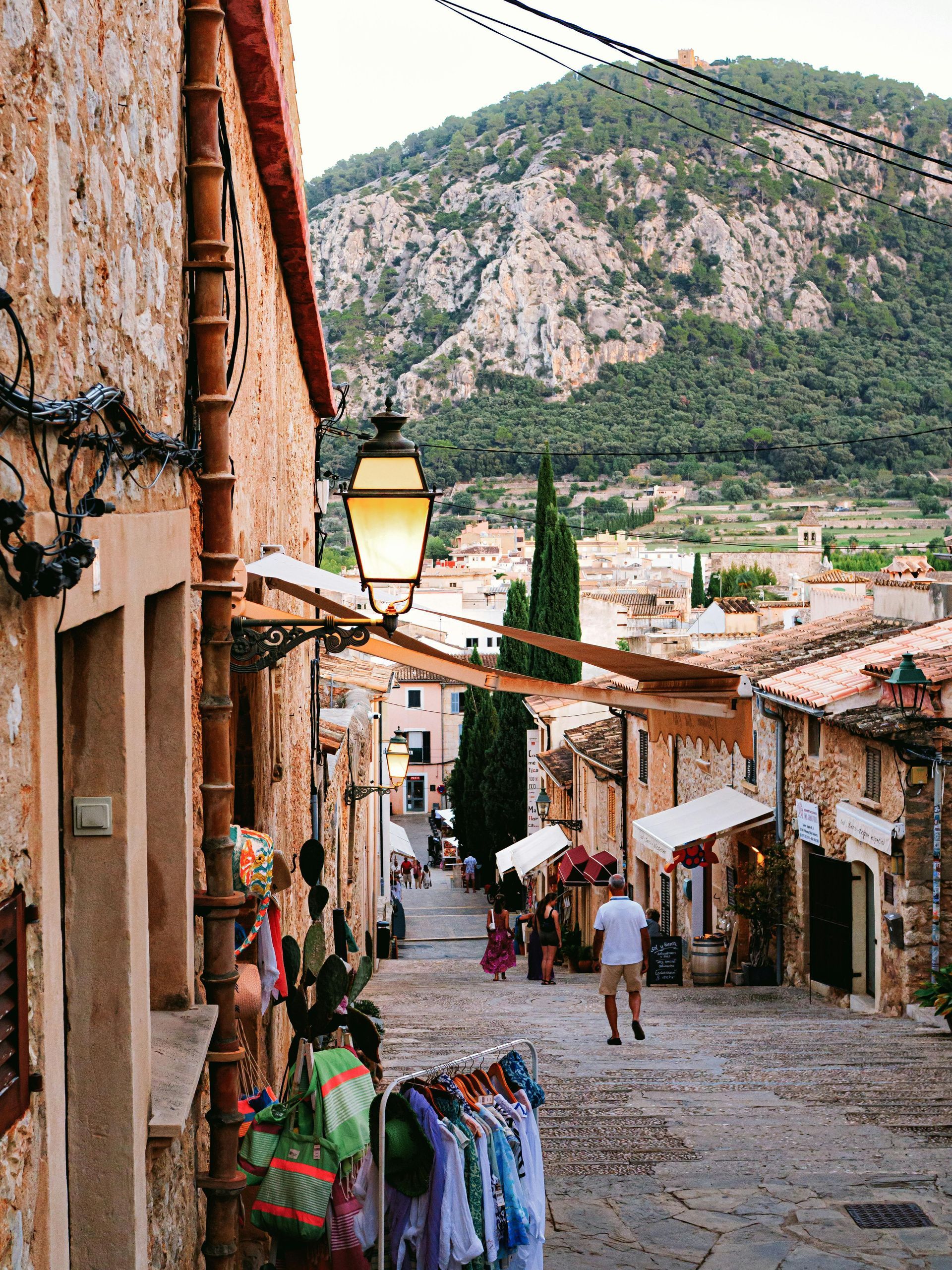 Stone street with shops, lantern, and mountain backdrop. People walk the cobblestone path.