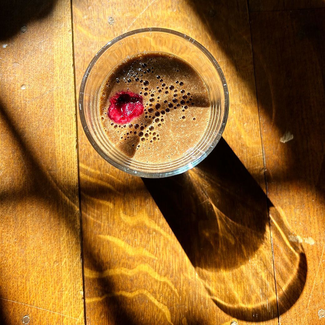Glass of drink with raspberry on a wooden surface, casting a long shadow in sunlight.