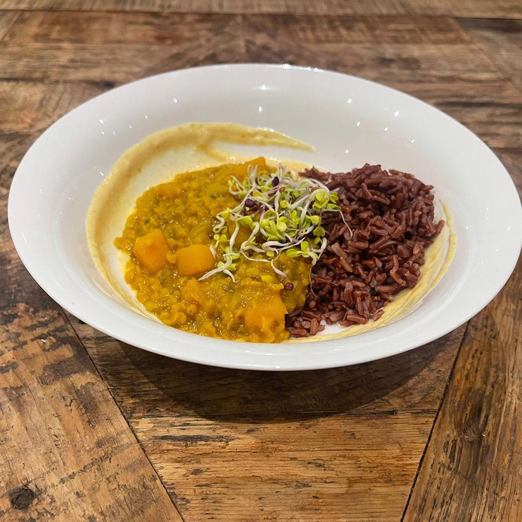 Bowl of food: yellow curry with red rice, sprouts, and creamy sauce. Wooden table.