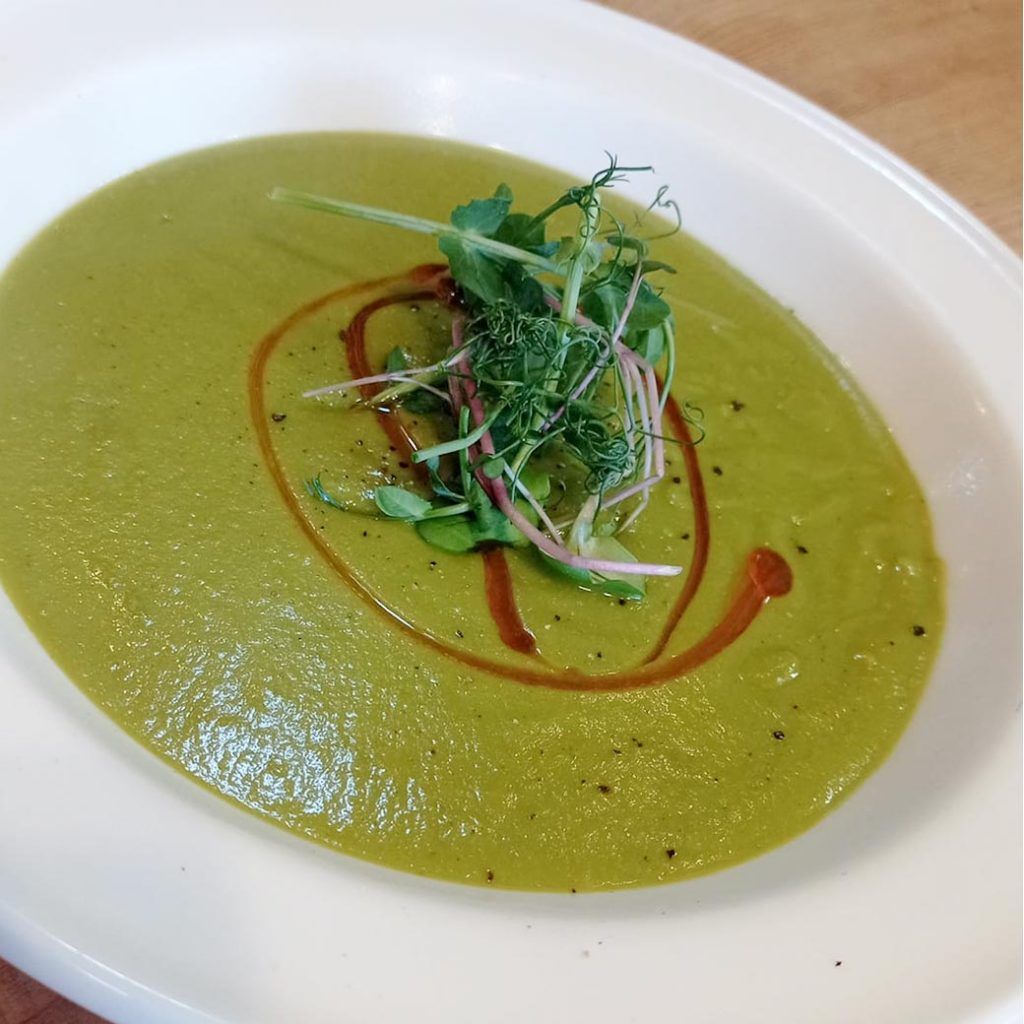 Green soup in white bowl, garnished with greens and balsamic glaze.
