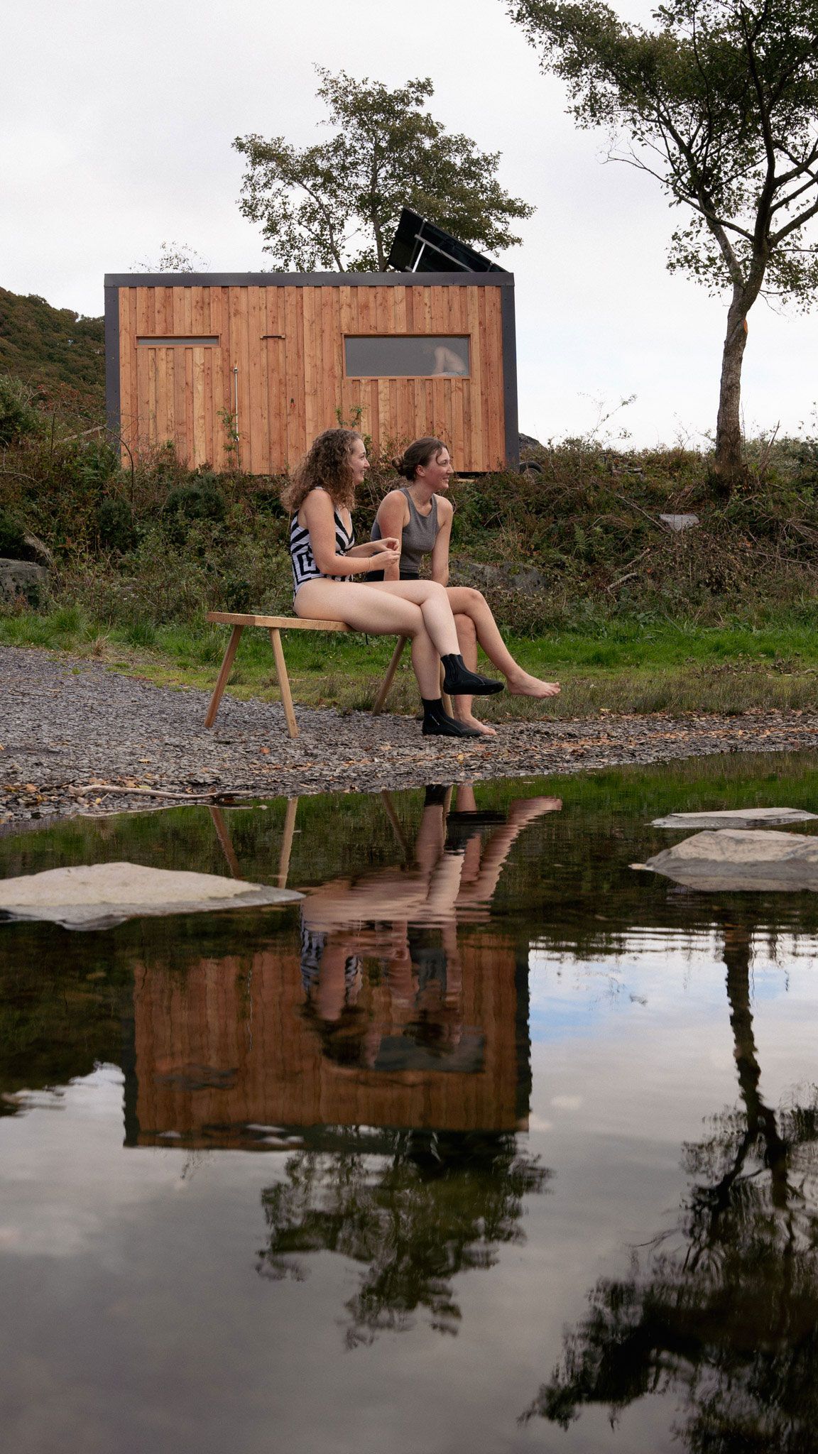 Two people sit on a bench near a river, reflecting a small wooden building in the water.