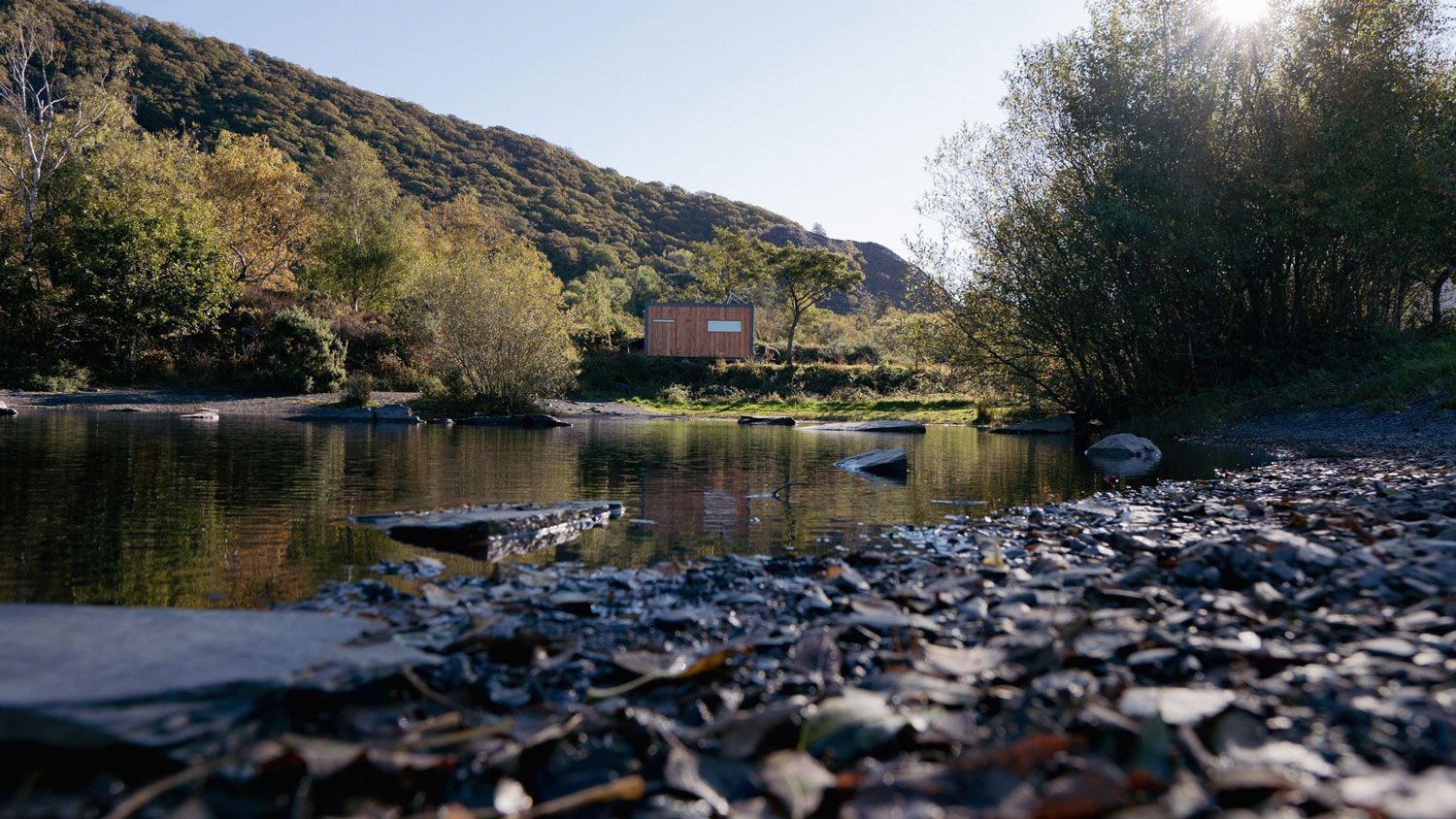 A serene river scene with a small building nestled near a tree-covered hillside on a sunny day.