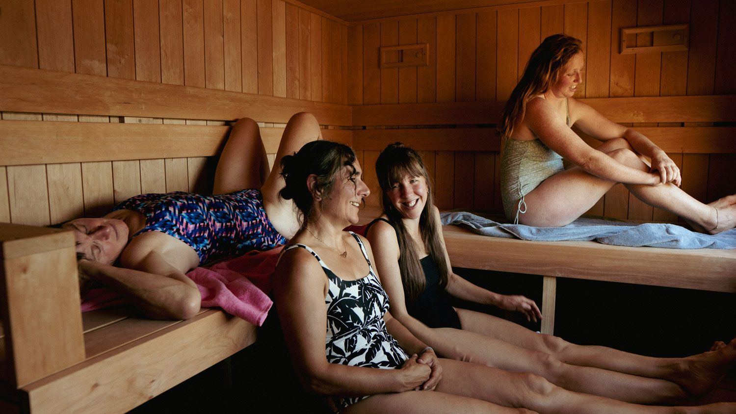 Four people relaxing in a wooden sauna, wearing swimsuits, smiling and chatting.