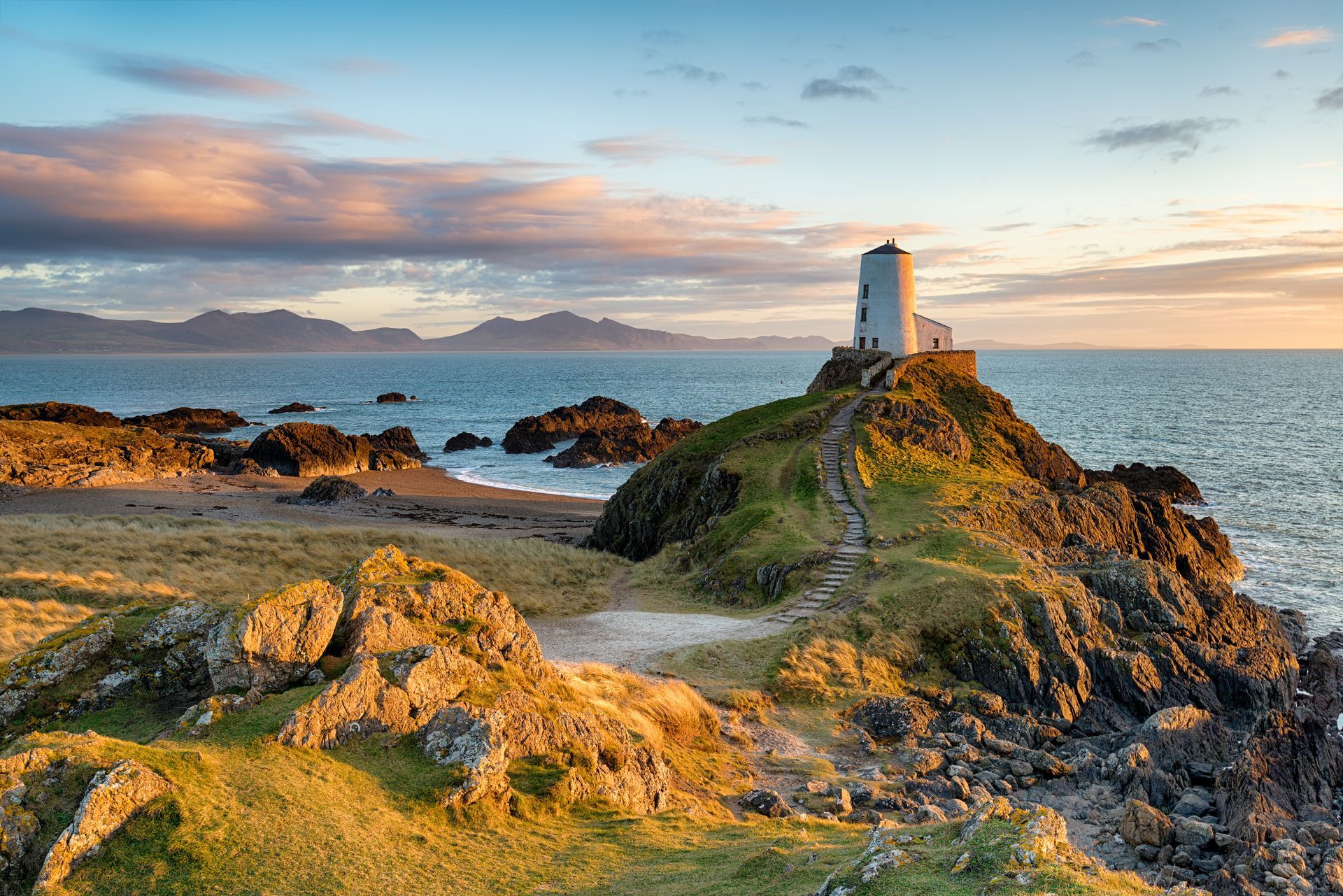 Lighthouse on rocky island at sunset, golden light, ocean, mountains in distance.