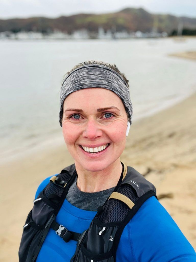 Woman smiling on a beach, wearing a headband, earbuds, and running vest. The ocean and trees are in the background.