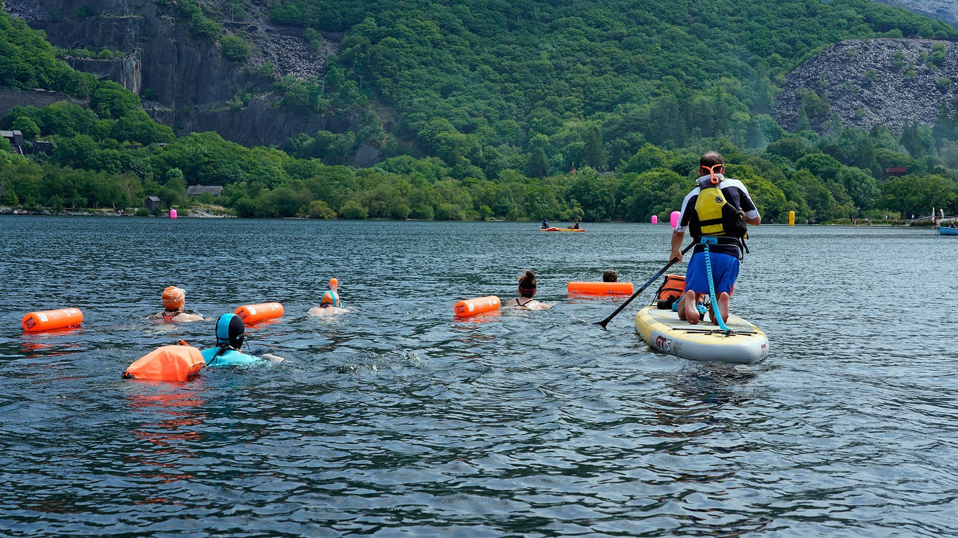 People swimming and paddleboarding in lake with mountain backdrop. Orange buoys mark a swim course.