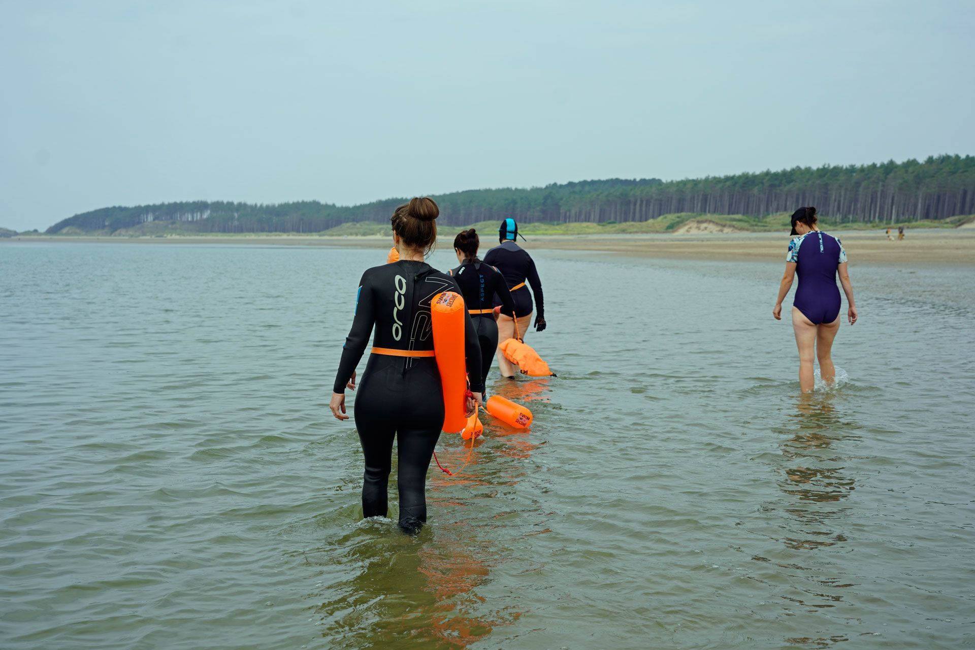 Four people wading into the ocean, wearing wetsuits, carrying orange buoys, beach in the background.