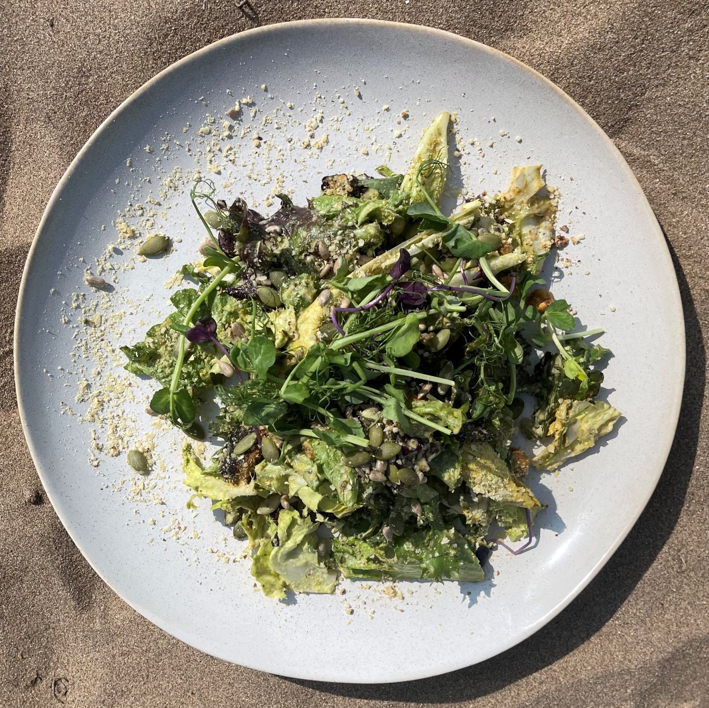 A green salad with seeds and sprouts on a plate, set on a sandy surface.