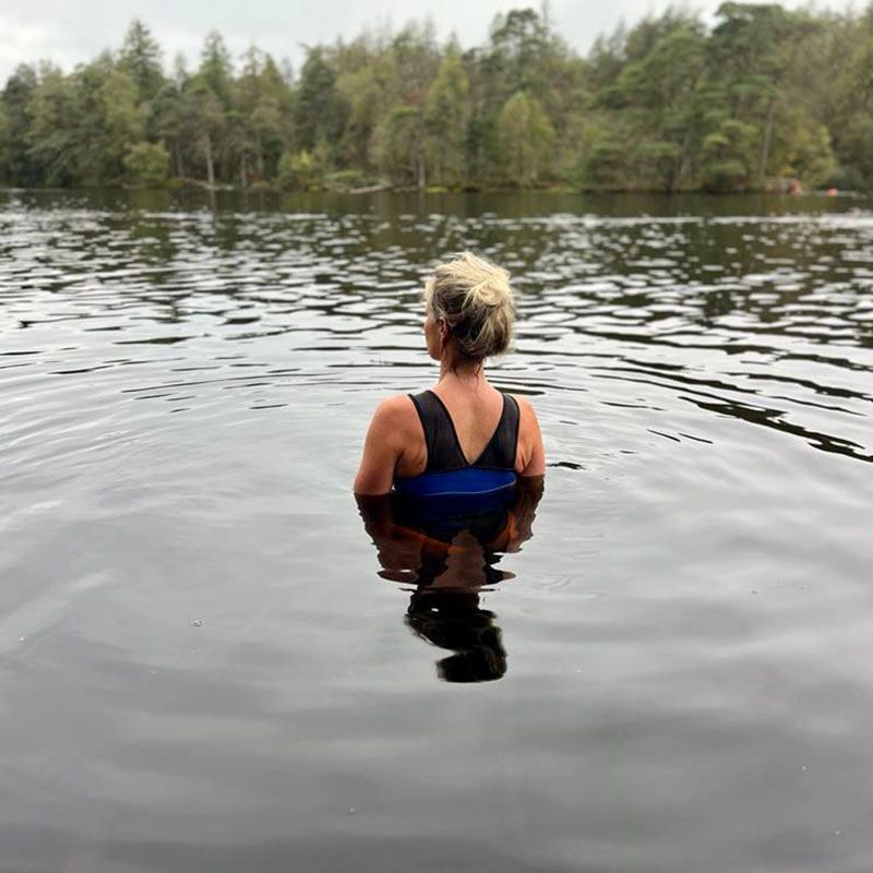 Woman in a black swimsuit stands in a lake, facing away from the camera. Trees line the far shore.
