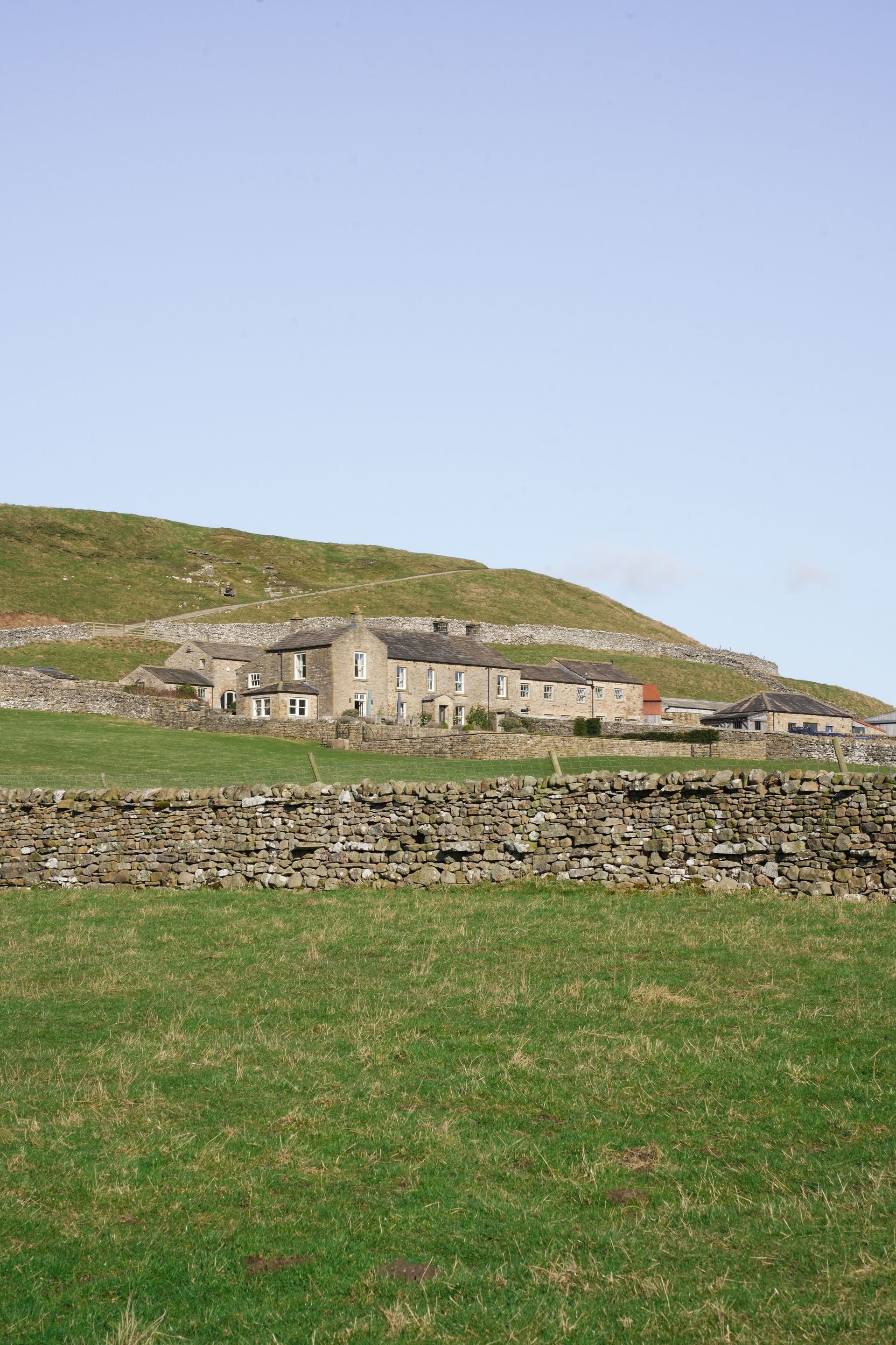 Stone cottages on a grassy hillside under a clear blue sky, with a low stone wall in the foreground