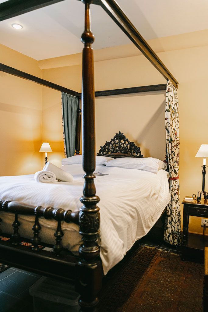 Bedroom with a dark wooden four-poster bed, white linens, and bedside lamps. Beige walls and patterned curtains.