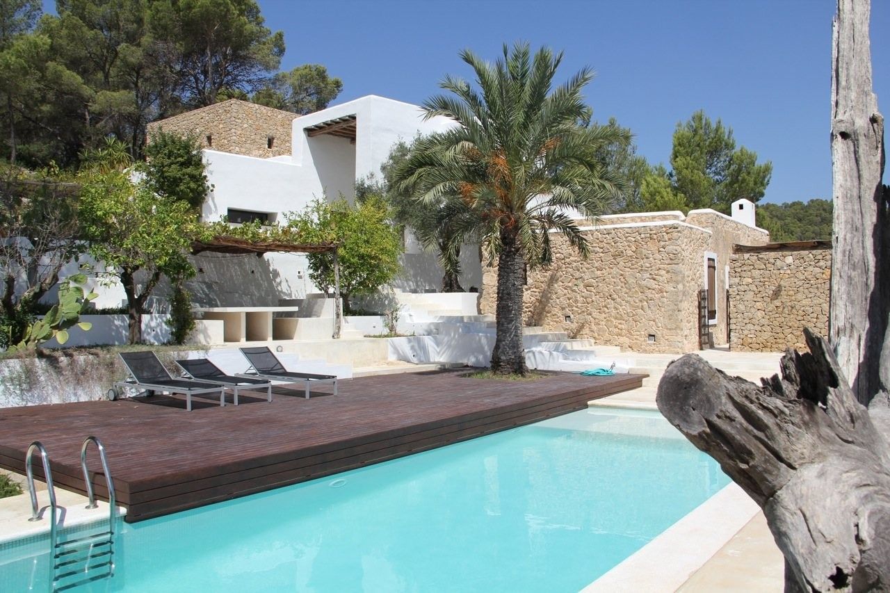 The pool and decked terrace below the whitewashed finca and stone houses, with a palm tree, Ibiza