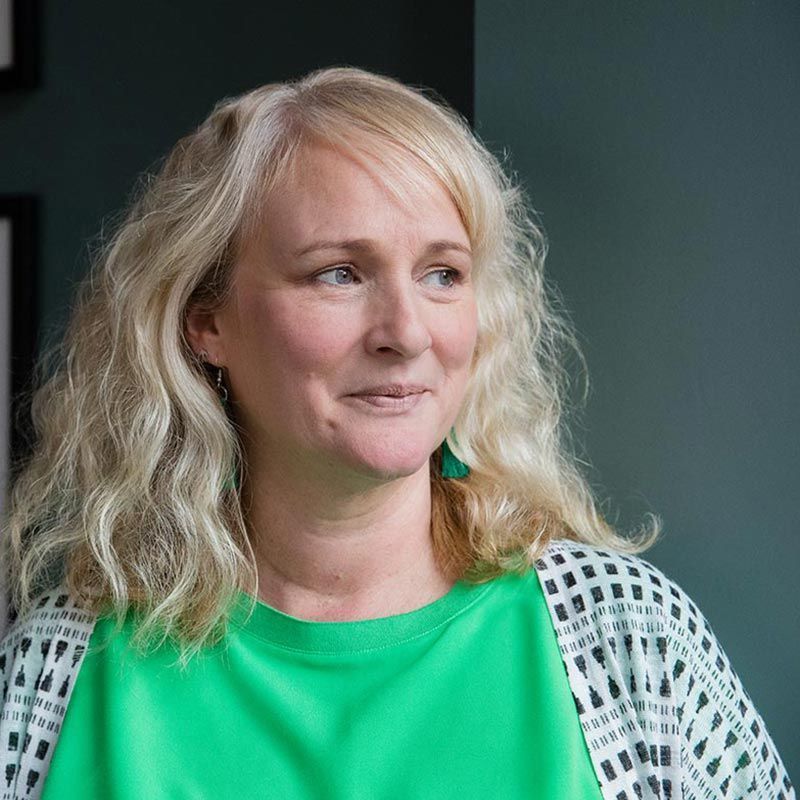 Blonde woman in green shirt and patterned cardigan, smiling, looking away, against a teal wall.