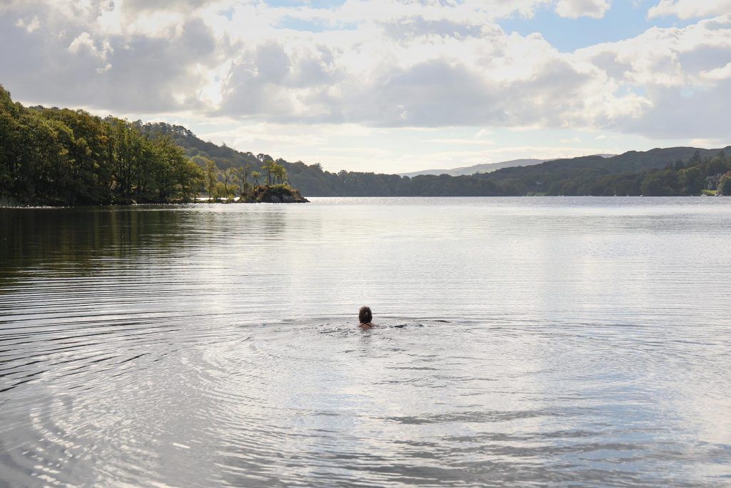 Person swimming in a calm lake towards distant tree-lined shores under a cloudy sky.