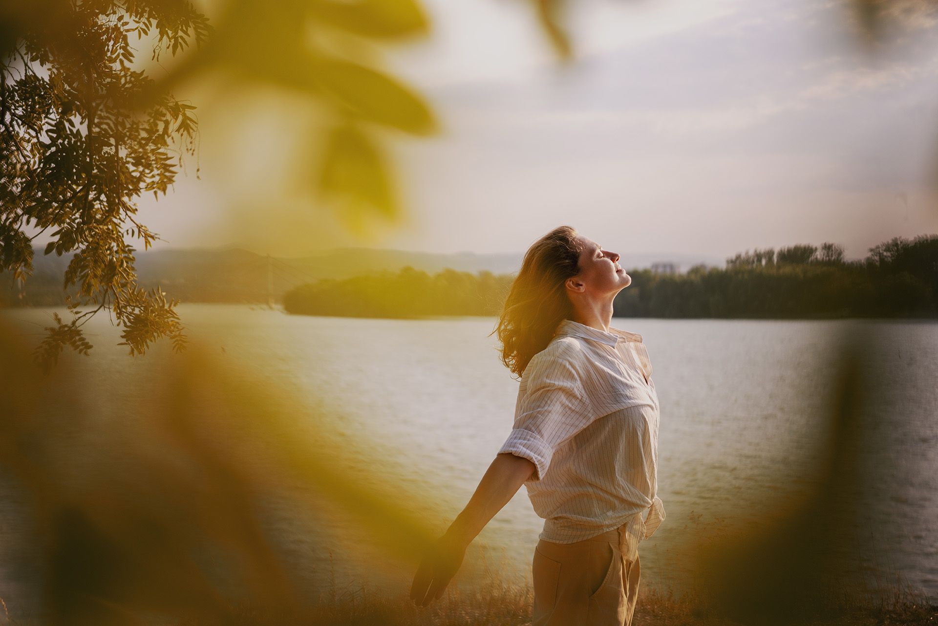 Woman stands by a lake, arms outstretched, eyes closed, enjoying the sunlight.