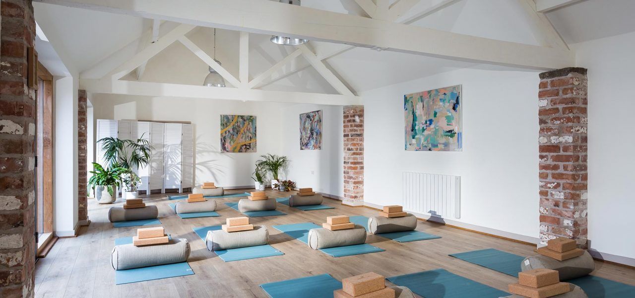 Yoga studio with mats, blocks, and bolsters on a wooden floor, brick columns, and white walls.