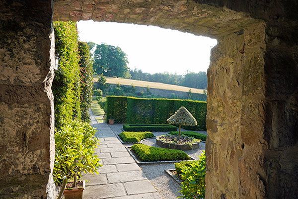 Stone archway frames a formal garden with hedges, walkways, and a tree, leading to a grassy hill.