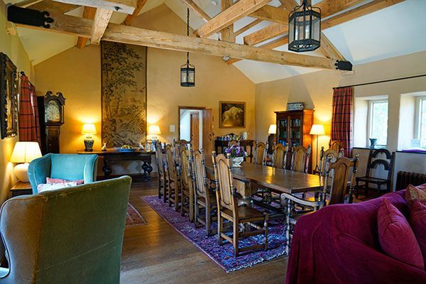 Long dining room with a large wooden table, chairs, and hanging lanterns. Beige walls, wooden beams, and a purple rug.