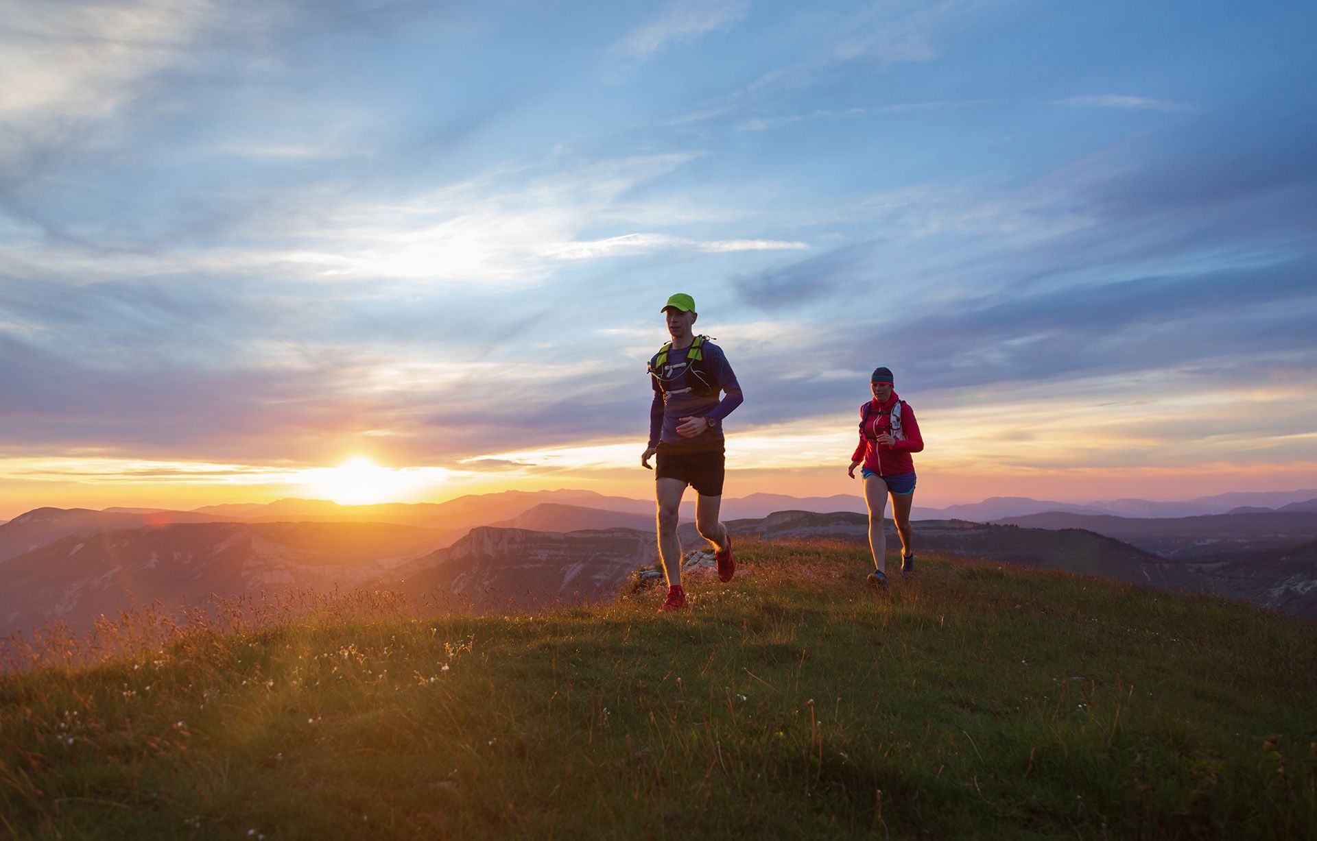 Two runners on a grassy ridge at sunset. Hills in the distance, blue sky with orange and yellow hues.