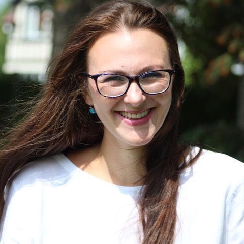 Woman with long brown hair, wearing glasses, and smiling outdoors.