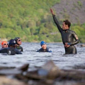 Man in wetsuit raises arm, instructing swimmers in a lake.