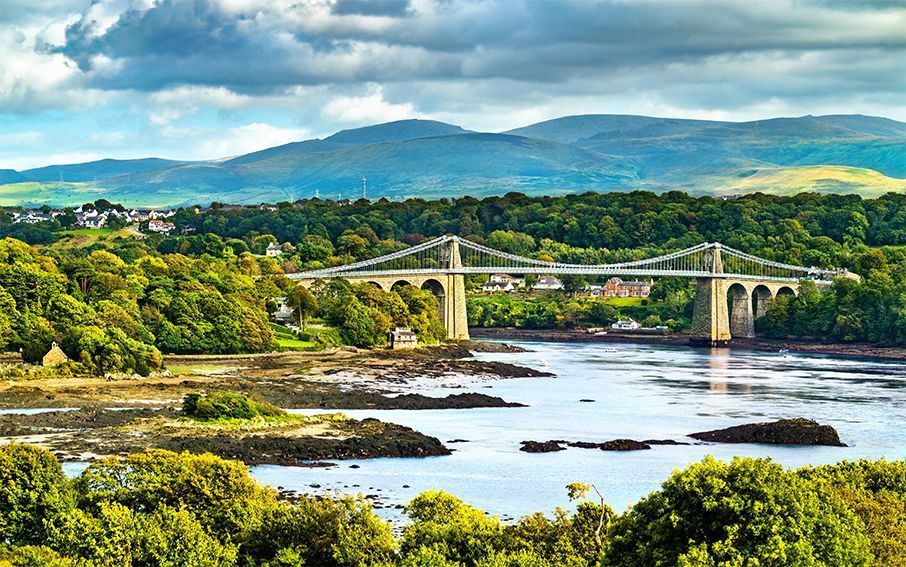 Bridge over water with green trees and mountains in the background under a cloudy sky.