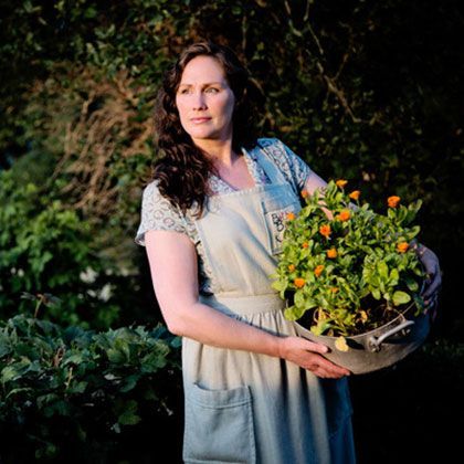 Woman in apron holds a pot of orange flowers in a garden, looking off-camera.