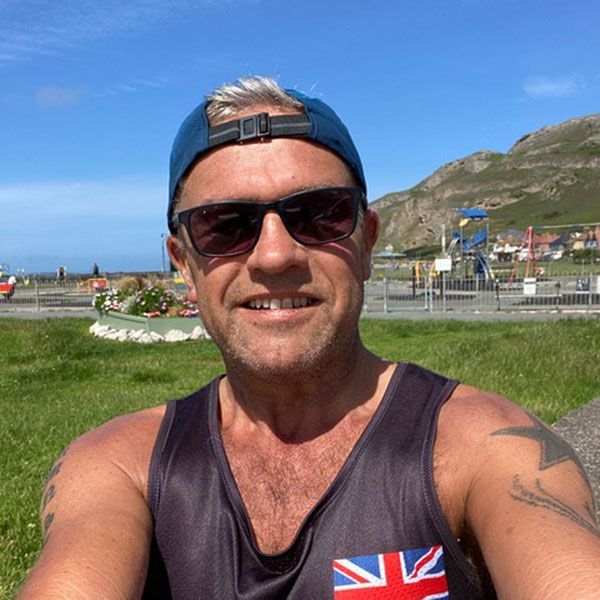 Man in sunglasses and hat smiles outdoors near water, with Union Jack patch on tank top.
